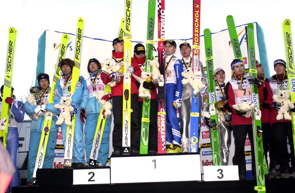 Łukasz Kruczek wraz: Adamem Małyszem, Robertem Mateją i Wojciechem Skupniem na podium konkursu drużynowego PŚ w skokach w Villach (09.12.2001) Zwycięzcy konkursu drużynowego w skokach narciarskich stoją na podium z nartami i maskotkami, uśmiechnięci sportowcy w zimowych strojach prezentują medale, tło stanowią banery sponsorów.
