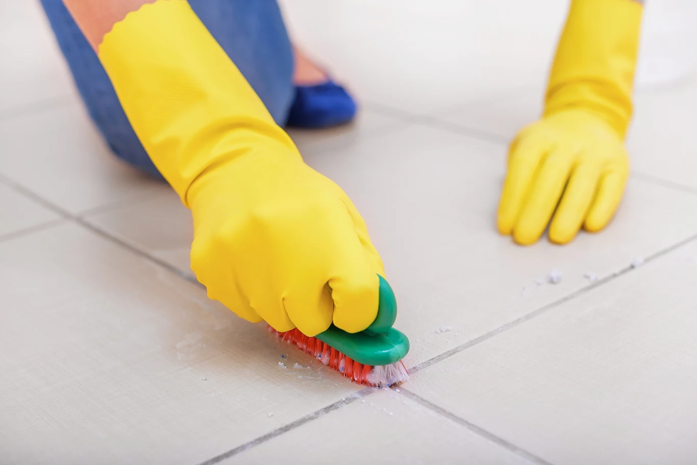 Ręce w żółtych rękawiczkach czyszczą szczotką fugi między płytkami na jasnej, ceramicznej podłodze. Widoczne drobinki zabrudzeń oraz dbałość o dokładność sprzątania.