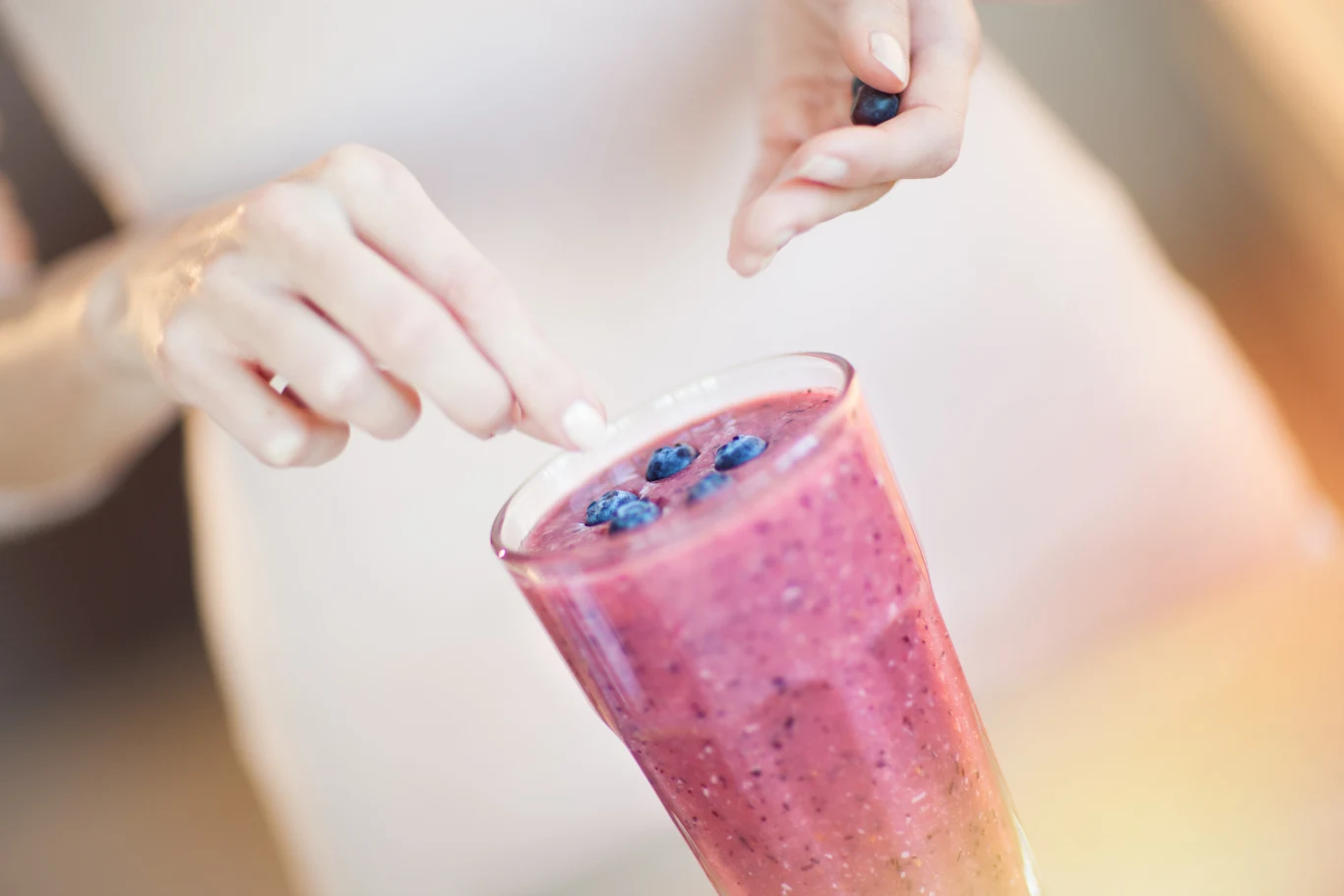 Koktajle to szybki, smaczny i realny sposób, aby skóra wyglądała młodziej: bardziej sprężyście, równomiernie i świetliście Szklanka wypełniona różowym smoothie udekorowana świeżymi jagodami, na pierwszym planie widoczne dłonie osoby dodającej owoce.