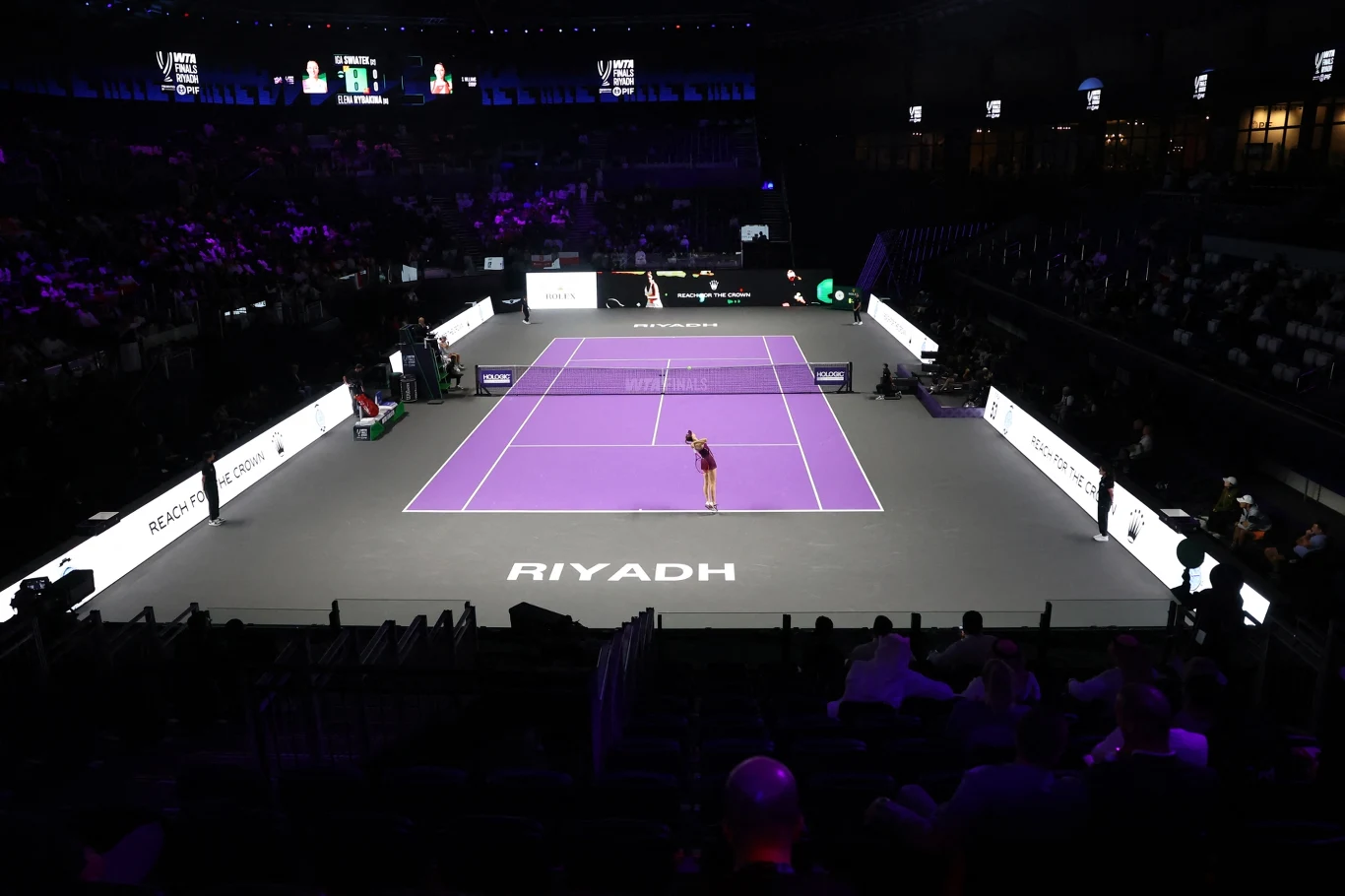 Główny kort WTA Finals 2025 w Rijadzie Nowoczesny kort tenisowy oświetlony sztucznym światłem, z widownią w tle i jednym zawodnikiem stojącym na boisku. Na podłożu widoczne duże napisy 'Riyadh', a otoczenie uzupełniają elektroniczne bandy reklamowe.
