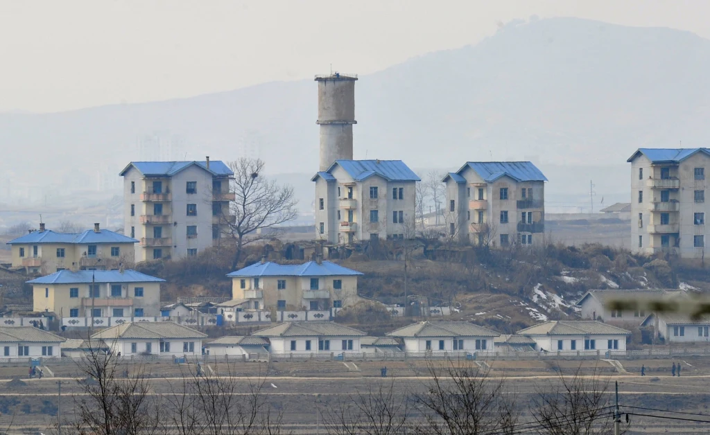 Kijong-Dong to obecnie "miasto duchów" Zabudowa mieszkalna z charakterystycznymi budynkami o niebieskich dachach, centralnie widoczna wieża. Teren otoczony pustymi polami i wzgórzami w tle.