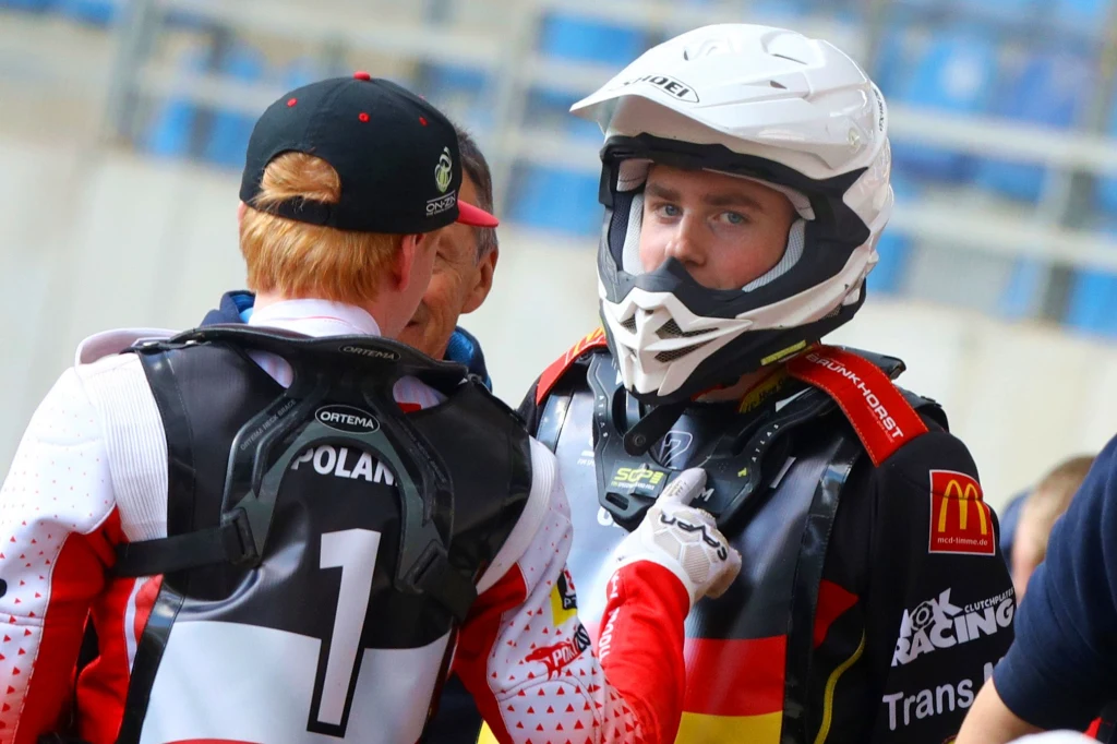 Dwóch motocyklistów ubranych w stroje wyścigowe i kaski rozmawia ze sobą przed rozpoczęciem zawodów na trybunach stadionu, z widocznymi oznaczeniami drużyn na ubraniach.