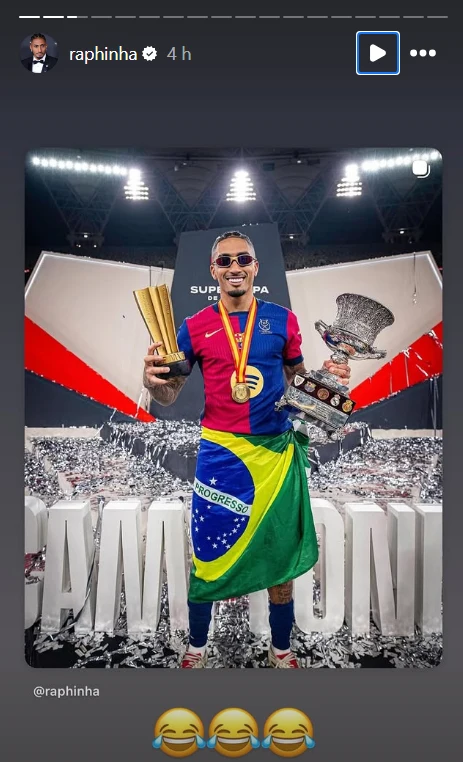 Sportowiec ubrany w kolorową koszulkę klubu piłkarskiego i owinięty flagą Brazylii trzyma w rękach dwa puchary i ma medal na szyi, stoi na tle dużego transparentu z napisem „Campeones” oraz trybun stadionu, wokół rozrzucone konfetti.