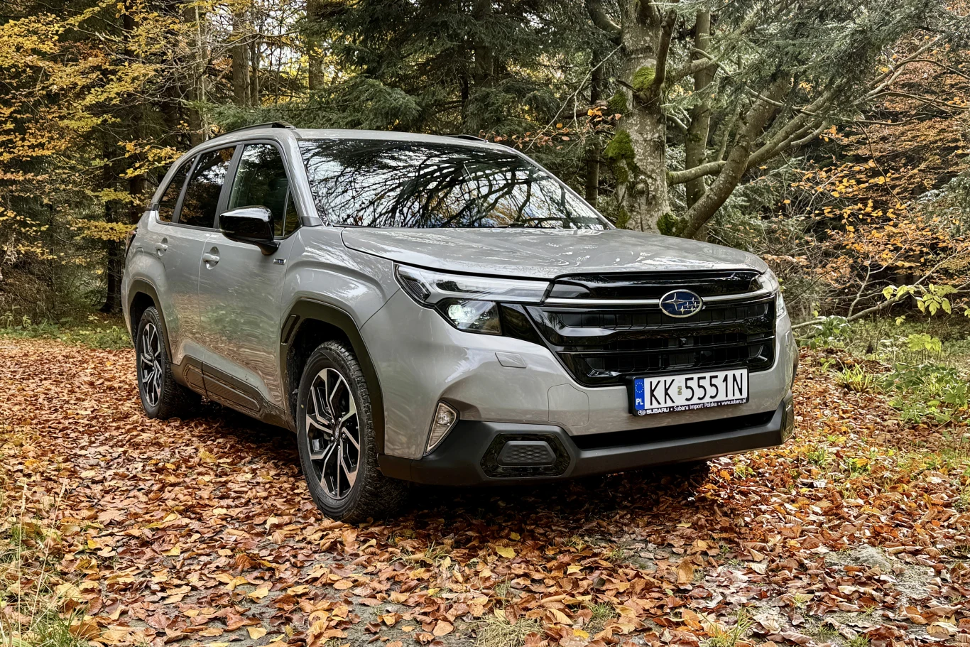 Srebrny samochód Subaru Forester stoi na leśnej drodze pokrytej opadłymi liśćmi, w otoczeniu drzew z jesiennymi barwami.