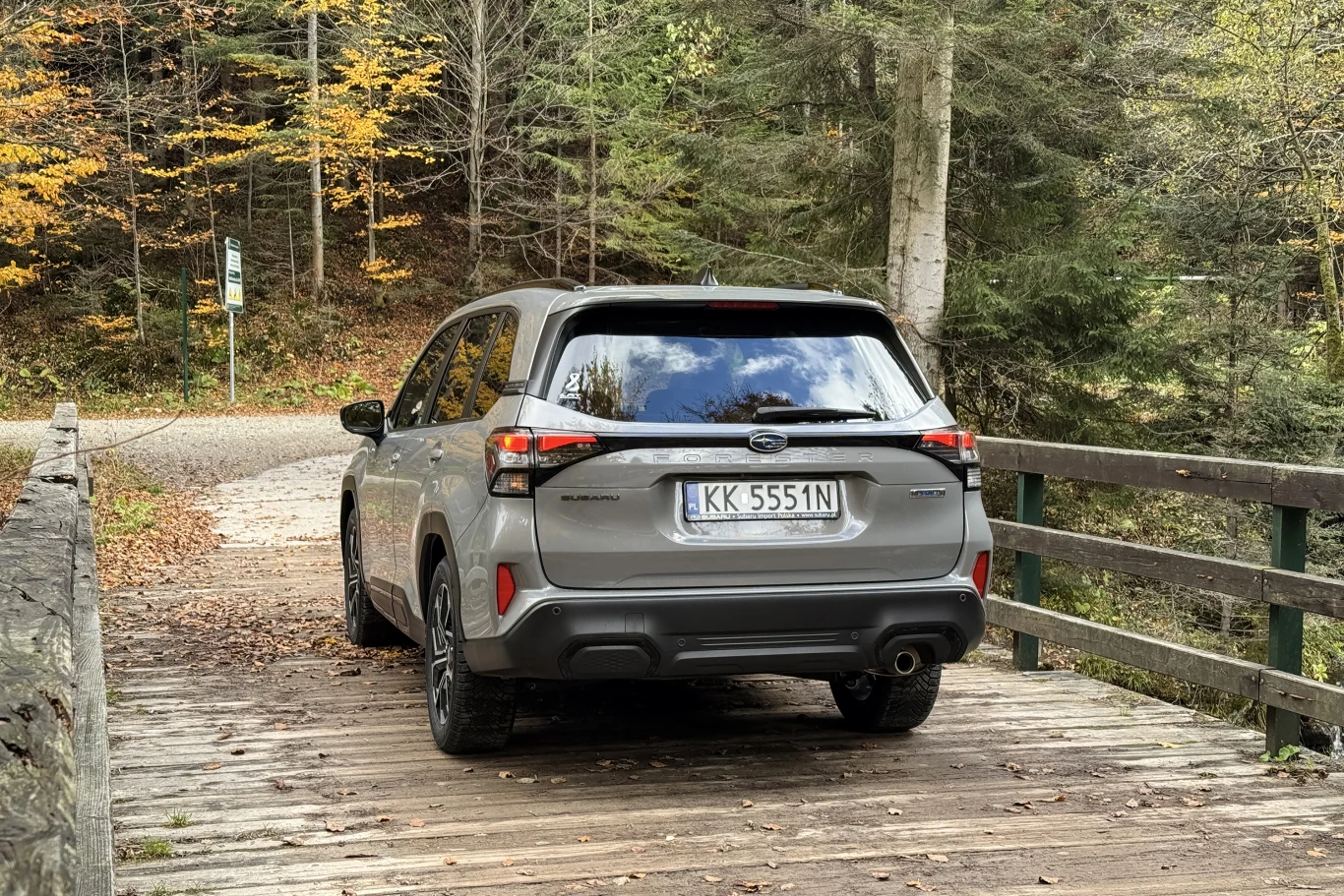 Samochód osobowy na drewnianym moście pośród leśnej scenerii z jesiennymi liśćmi. W tle widoczne drzewa iglaste i liściaste oraz fragment asfaltowej drogi.