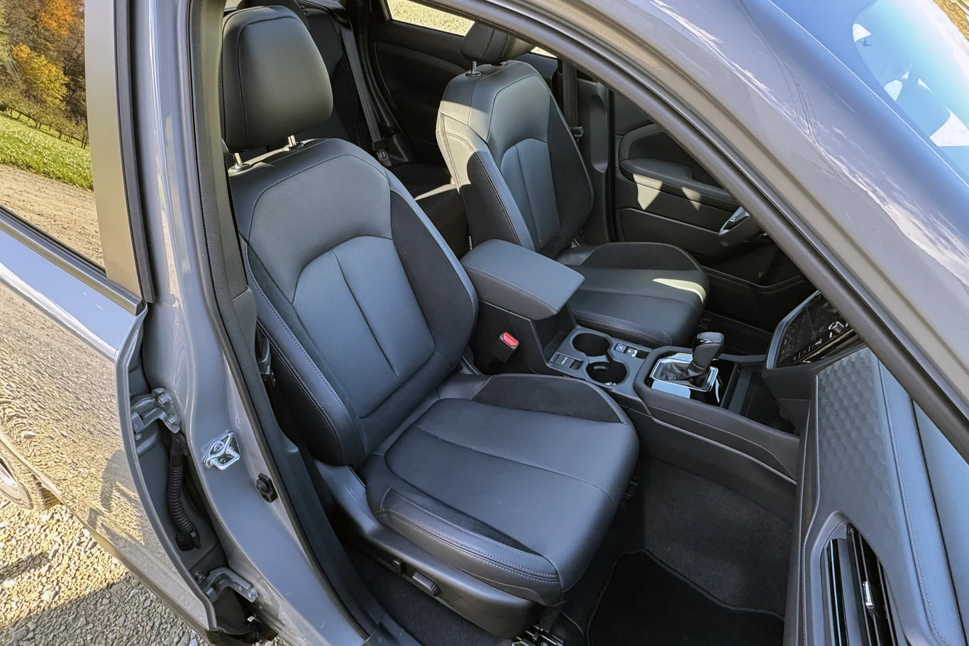 Wnętrze samochodu z widocznymi dwoma przednimi fotelami wykonanymi z ciemnej skóry, centralnym podłokietnikiem oraz konsolą środkową z uchwytami na napoje i elementami sterującymi.