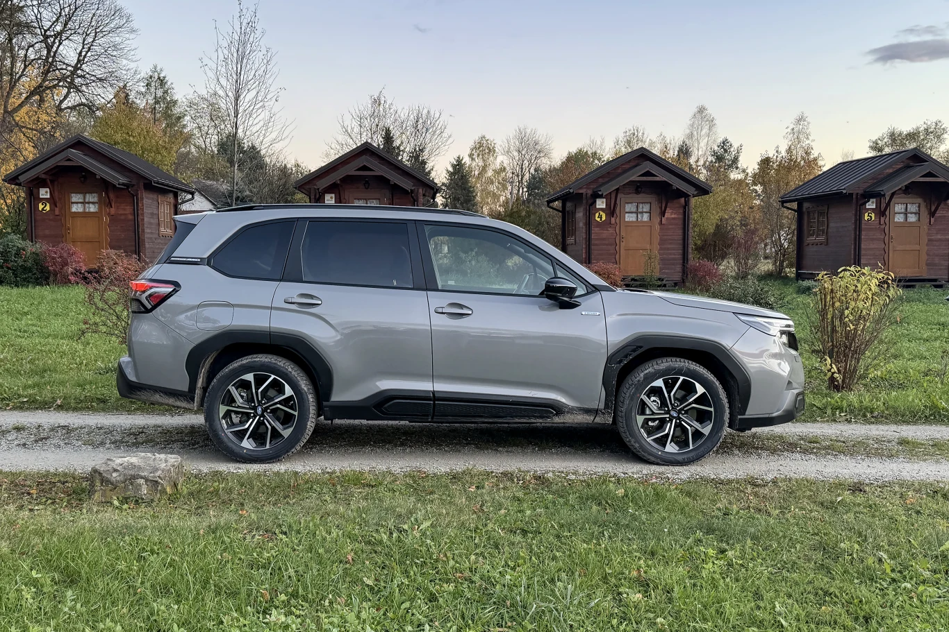 Srebrny samochód typu SUV stoi na żwirowej drodze, w tle widoczne są cztery drewniane domki letniskowe otoczone zielenią oraz jesiennymi drzewami.