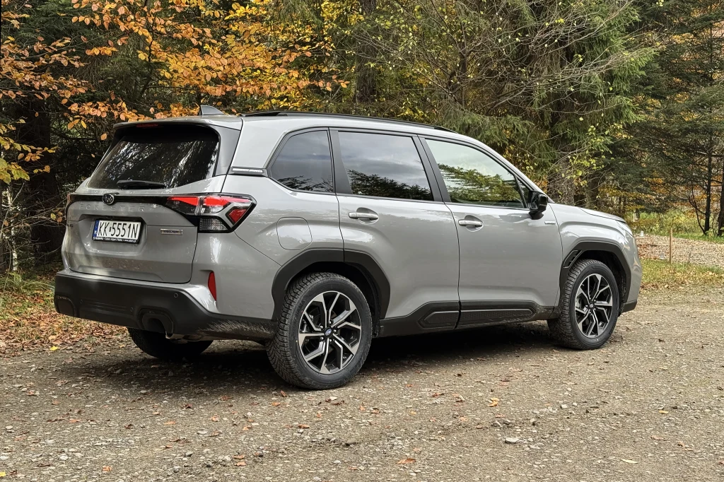 Srebrny SUV marki Toyota Corolla Cross zaparkowany na leśnej drodze żwirowej, otoczony jesiennymi drzewami i bujną roślinnością w tle.