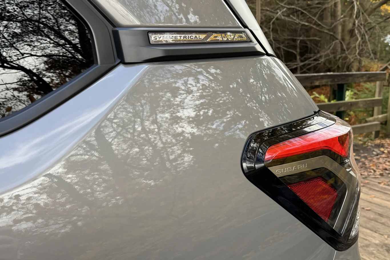 Tylna część srebrnego samochodu z widocznym światłem tylnym oraz napisem 'Symmetrical AWD', w tle fragment drewnianego płotu i drzewa.
