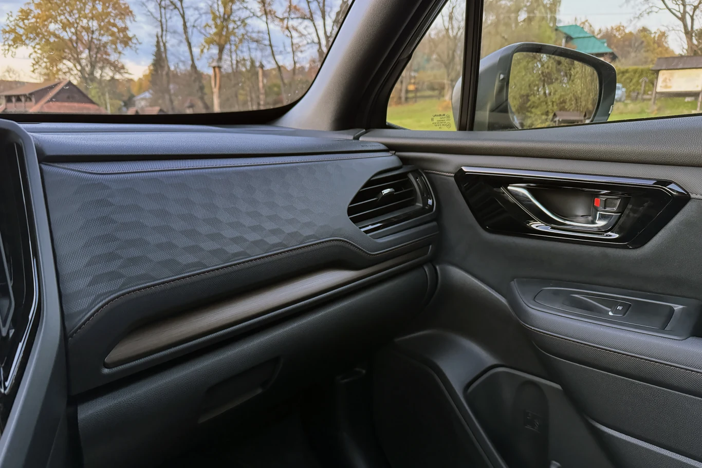 Wnętrze nowoczesnego samochodu z widocznym fotelem pasażera, wykończoną deską rozdzielczą, kratką wentylacyjną oraz bocznym lusterkiem za szybą. W tle przez okno widoczne jesienne drzewa oraz budynki na wsi.