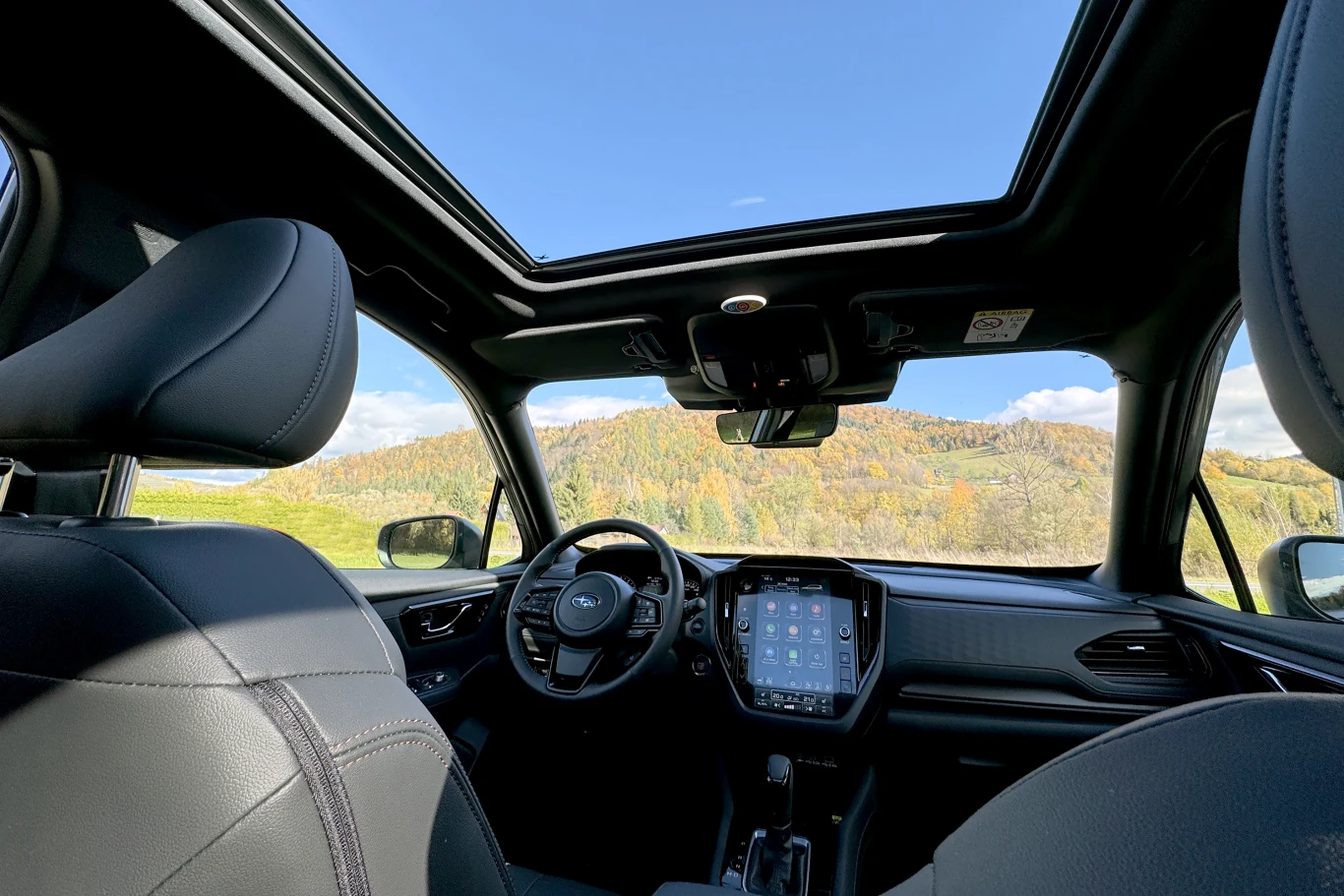 Wnętrze nowoczesnego samochodu z przeszklonym dachem, ekranem dotykowym na konsoli centralnej i wygodnymi fotelami, z widokiem na górzysty krajobraz za szybą.