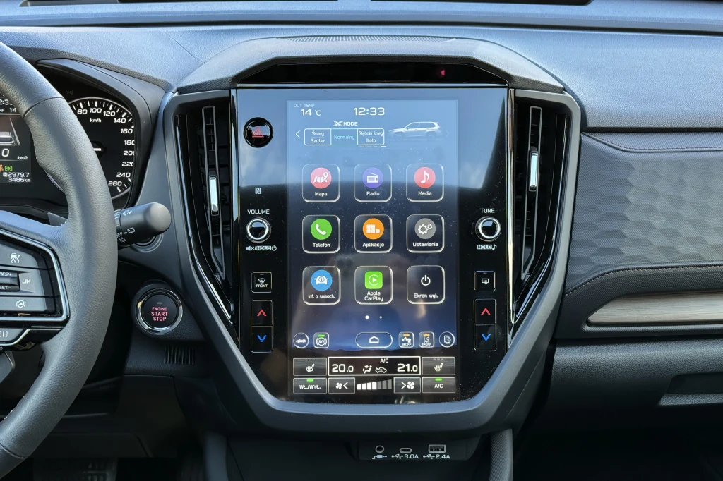 Duży dotykowy ekran centralny umieszczony w nowoczesnym wnętrzu samochodu, z wyświetlonym interfejsem multimedialnym, przyciskami sterującymi oraz panelami klimatyzacji, wokół widać fragmenty deski rozdzielczej i kierownicy.