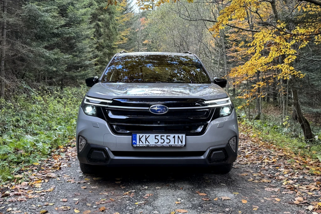 Szary samochód terenowy marki Subaru z widoczną tablicą rejestracyjną, zaparkowany na leśnej drodze otoczonej gęstymi drzewami i opadłymi, żółtymi liśćmi.