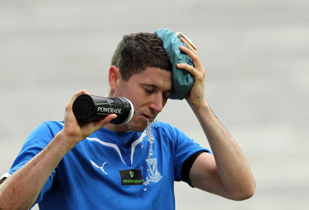 Robert Lewandowski w barwach Lecha Poznań Sportowiec w niebieskiej koszulce schładza się wodą z butelki oraz przykłada kompres do głowy, prawdopodobnie po wysiłku fizycznym.