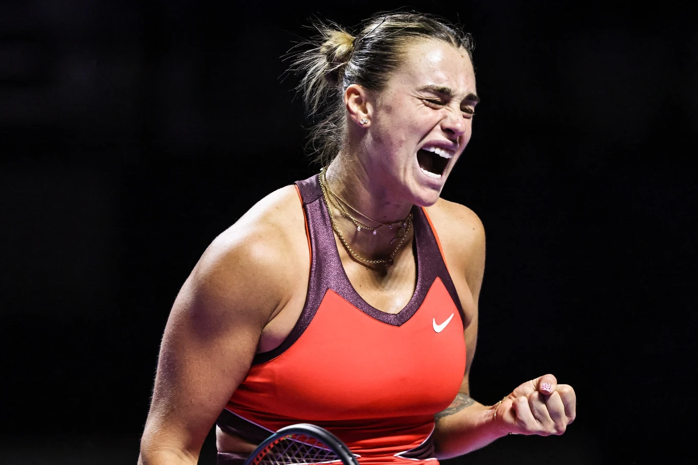 Aryna Sabalenka Zawodniczka tenisa w emocjonalnym momencie na korcie, ubrana w czerwono-fioletowy strój sportowy, ściska pięść i wykrzykuje z radości lub determinacji po udanej akcji.