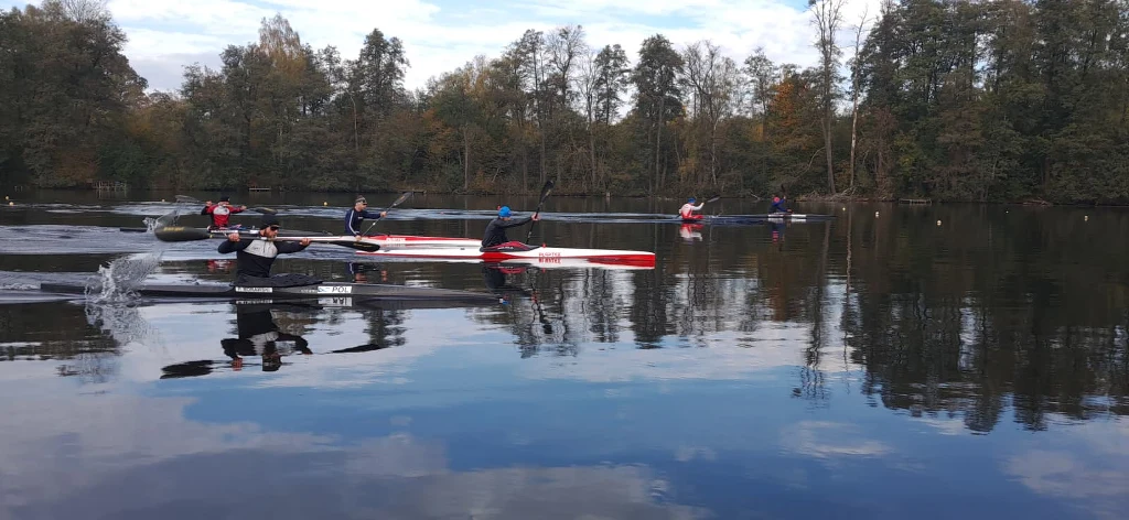 Grupa osób płynących kajakami i kanadyjkami po spokojnej wodzie, otoczenie stanowią drzewa w jesiennych barwach odbijające się na tafli jeziora.