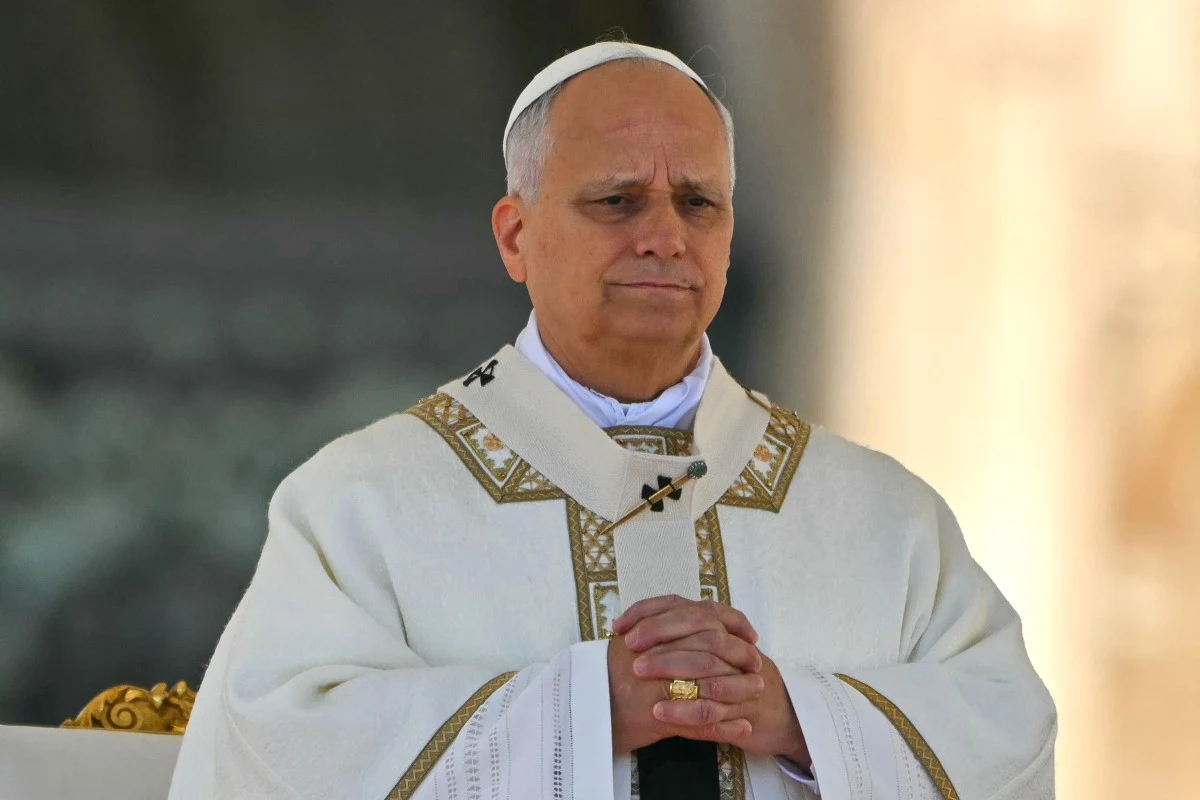 Papież Leon XIV Papież Leon XIV w białych szatach liturgicznych i mitrze, stojący ze złożonymi dłońmi w kościele.