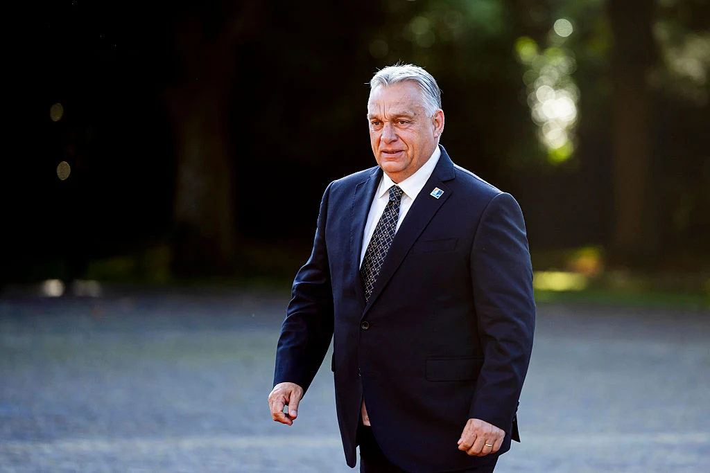 Premier Węgier Viktor Orban Mężczyzna w ciemnym, eleganckim garniturze oraz białej koszuli i krawacie stoi na otwartej przestrzeni, na tle rozmytych drzew i łagodnego światła.