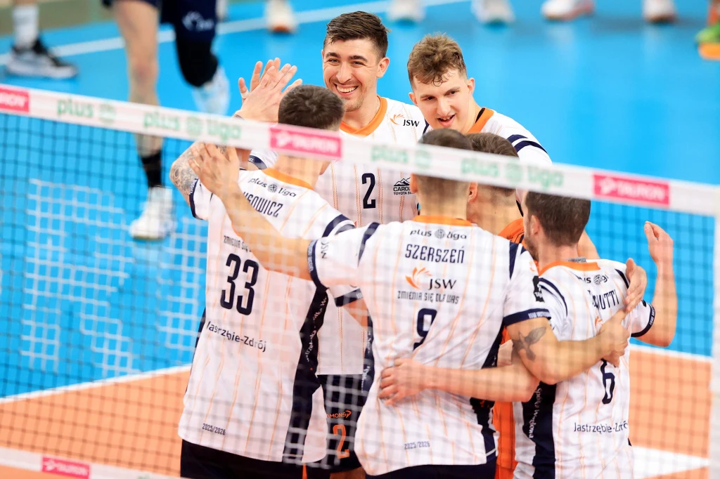 Grupa siatkarzy ubranych w jednolite stroje drużynowe świętują zdobycie punktu na parkiecie, obejmując się i przybijając piątki za siatką podczas meczu siatkarskiego.