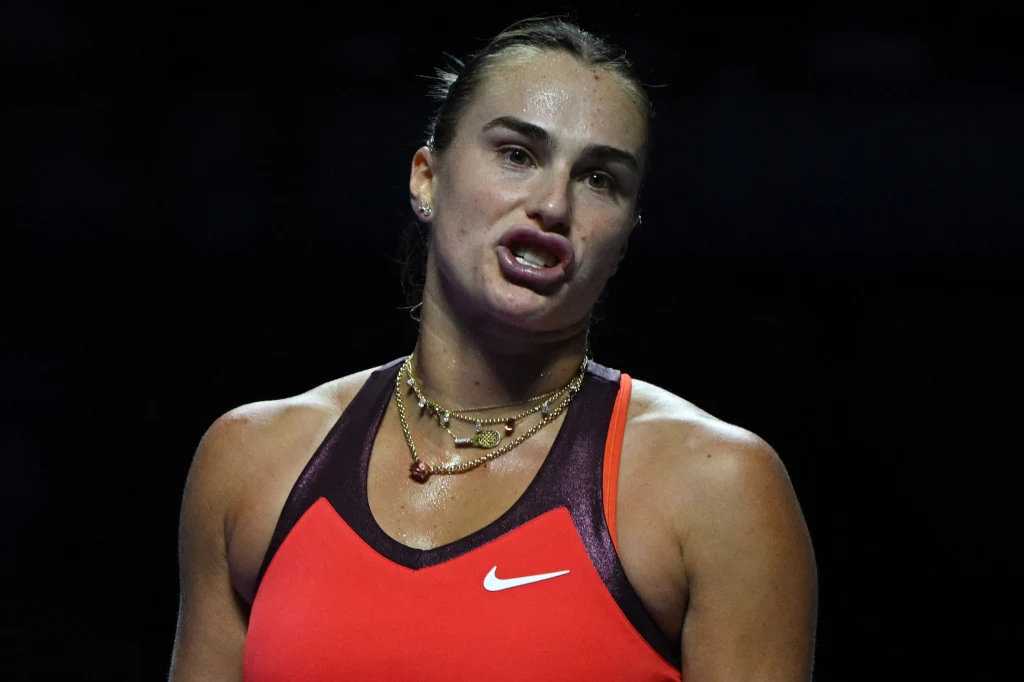 Aryna Sabalenka podczas WTA Finals 2025 Kobieta w sportowym stroju na tle ciemnego otoczenia, z wyrazem skupienia na twarzy, ubrana w czerwony top z widocznym logo Nike i kilka naszyjników na szyi.