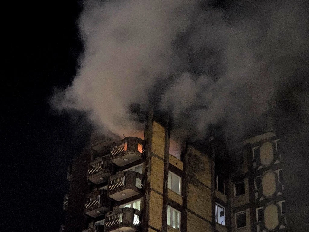 Pożar domu opieki dla seniorów w Tuzli Płomienie i gęsty dym wydobywające się z okien mieszkania w wysokim budynku nocą wskazują na trwający pożar. Nad balkonami rozprzestrzenia się ciemny dym, a światła w oknach poniżej wciąż się świecą.