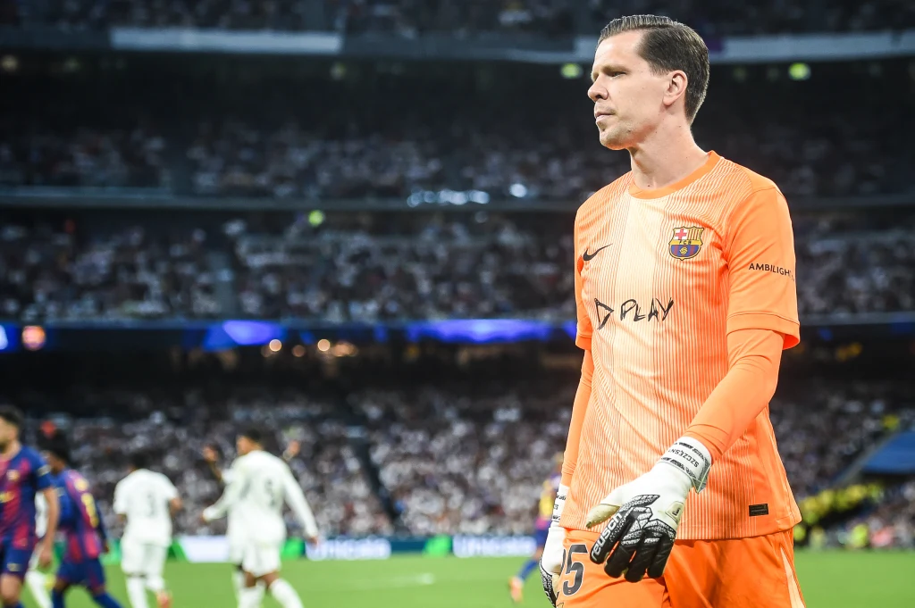 Bramkarz w pomarańczowym stroju z logo FC Barcelona na tle stadionu podczas meczu piłkarskiego, w oddali widoczni zawodnicy w białych i granatowych strojach, trybuny wypełnione kibicami.