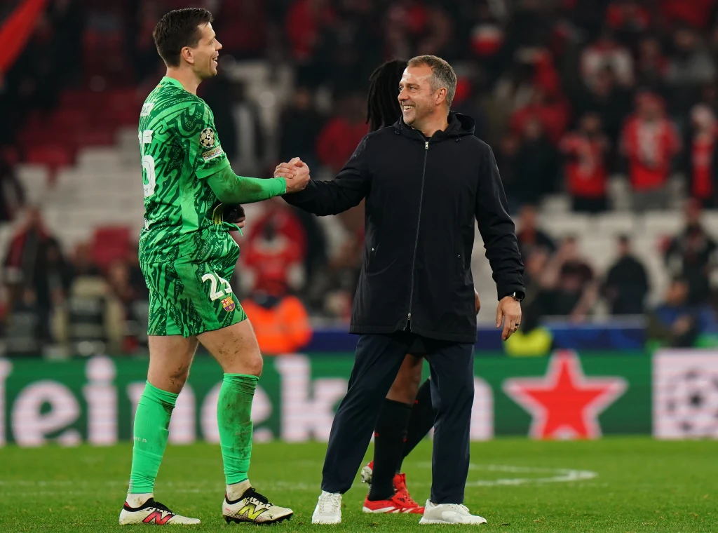 Wojciech Szczęsny i Hansi Flick Zawodnik w zielonym stroju piłkarskim wita się i uśmiecha do trenera stojącego obok niego na murawie stadionu. W tle rozmazana publiczność i elementy reklamowe.