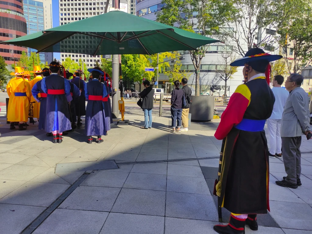 W Korei przy przejściach dla pieszych można zobaczyć parasole chroniące przed słońcem Grupa osób ubranych w barwne, tradycyjne stroje w centrum miasta, otoczona przez obserwatorów i nowoczesne budynki, całość oświetlona dziennym światłem.