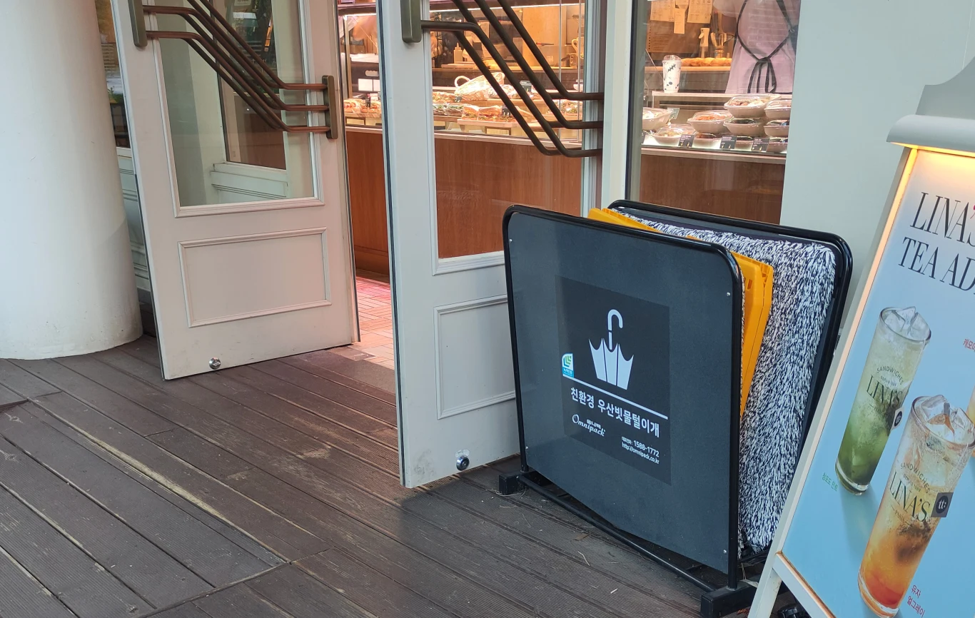 Otwór wejściowy do kawiarni lub piekarni z przeszklonymi drzwiami, stojak na parasole z oznaczeniem w języku koreańskim oraz fragment stojaka reklamującego napoje mrożone. W tle, za drzwiami, widoczne wypieki na półkach.