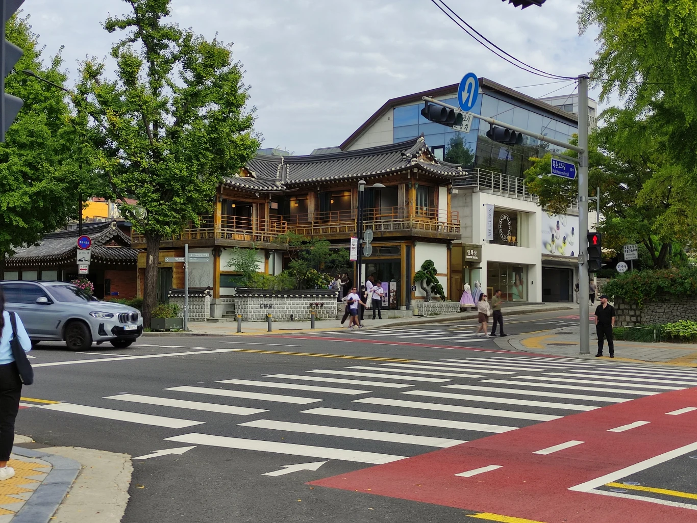 Skrzyżowanie ulic w mieście, z zebrą dla pieszych, samochodem zatrzymującym się przed przejściem oraz grupką pieszych przechodzących na drugą stronę. W tle klasyczna zabudowa azjatycka z drewnianym balkonem oraz nowoczesne budynki, otoczona zielenią dr...