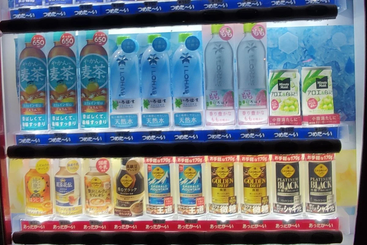 Automat z napojami oferujący różnorodne napoje butelkowane, w tym zielone herbaty, napoje izotoniczne oraz kawy w puszkach. Produkty ustawione są w trzech rzędach, widoczne japońskie etykiety.