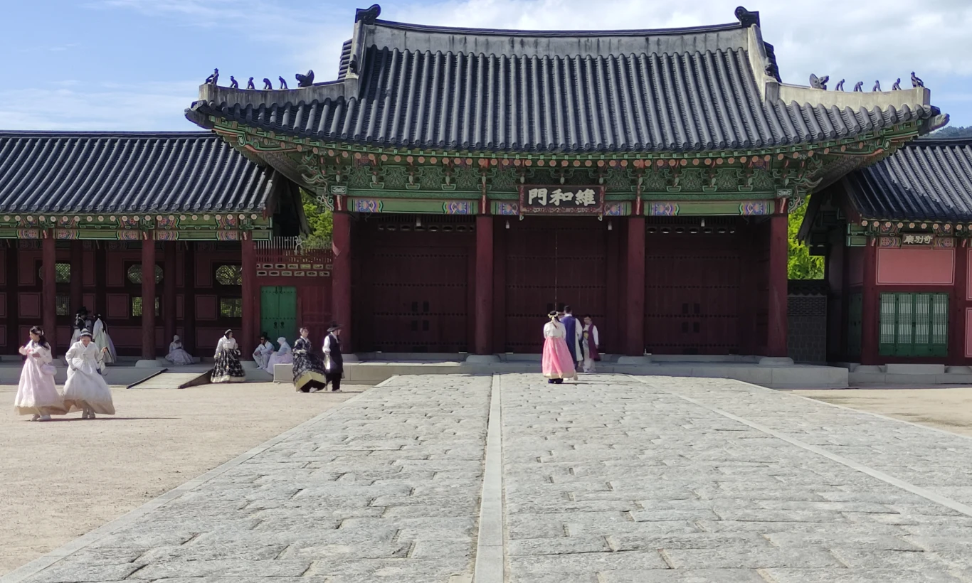 Tradycyjny pałac azjatycki z szeroką kamienną drogą prowadzącą do ozdobnej bramy. Przed budynkiem spacerują osoby ubrane w tradycyjne stroje, głównie kobiety w hanbokach. Słoneczna pogoda i niebieskie niebo dodają scenerii harmonii.