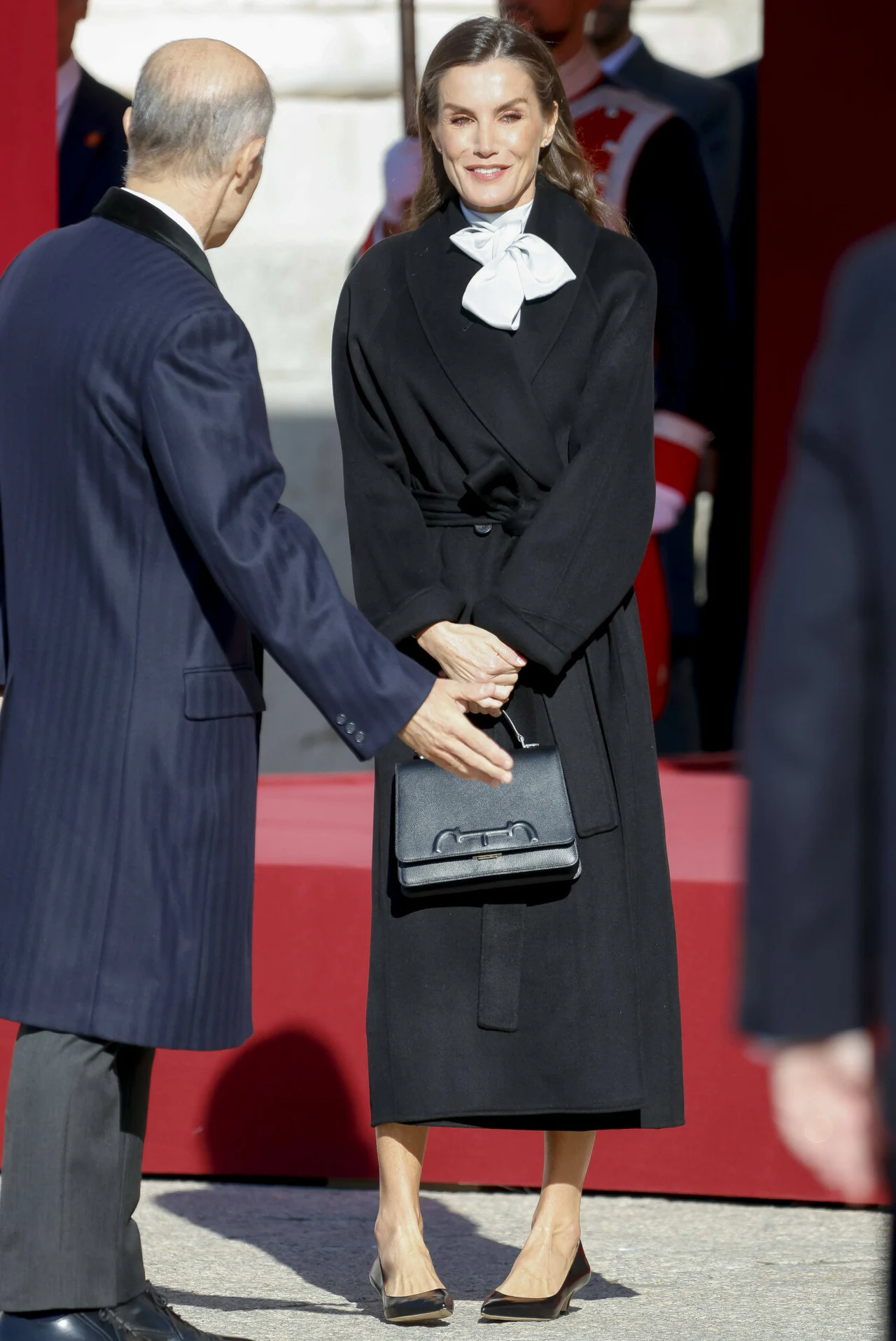 Płaszcz zachwycał szczegółami. To klasa sama w sobie Elegancko ubrana kobieta w czarnym płaszczu z białą kokardą na szyi stoi obok mężczyzny w garniturze, trzyma w rękach czarną torebkę, w tle czerwony podest i mundurowi.