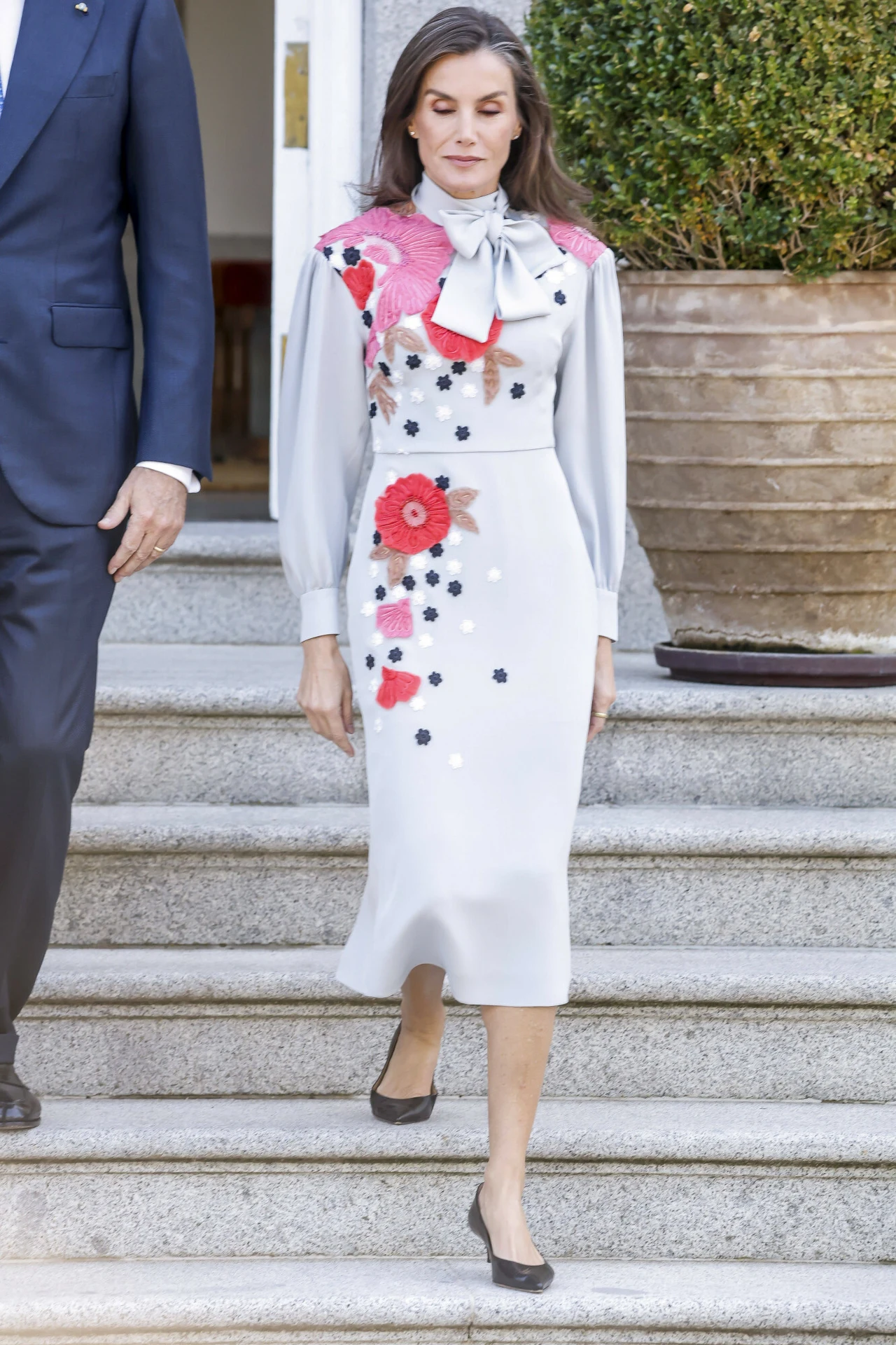 Sukienka, którą królowa skrywała pod płaszczem jest przeciwieństwem stonowanej czerni Kobieta w eleganckiej, jasnej sukience z ozdobnymi motywami kwiatowymi, schlektanym kołnierzykiem i kokardą na szyi schodzi po schodach w towarzystwie mężczyzny w ciemnym garniturze. W tle widać duży doniczkowy krzew.