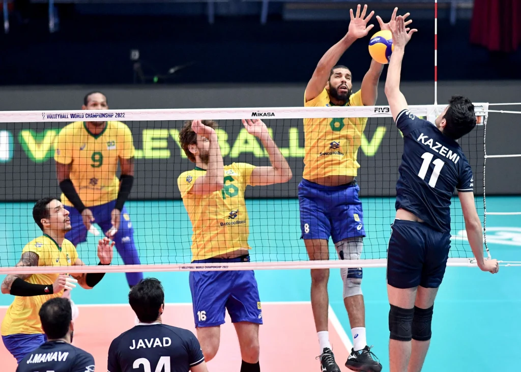 Trzech zawodników w żółtych koszulkach blokuje piłkę przy siatce podczas próby ataku siatkarza w granatowej koszulce z numerem 11. W tle widoczni są kolejni gracze i fragment boiska siatkarskiego.