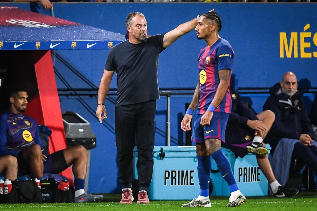 Hansi Flick i Raphinha Trener drużyny piłkarskiej delikatnie dotyka głowy zawodnika w stroju FC Barcelona, obok ławka rezerwowych oraz lodówki sponsora Prime, w tle trybuna stadionu.