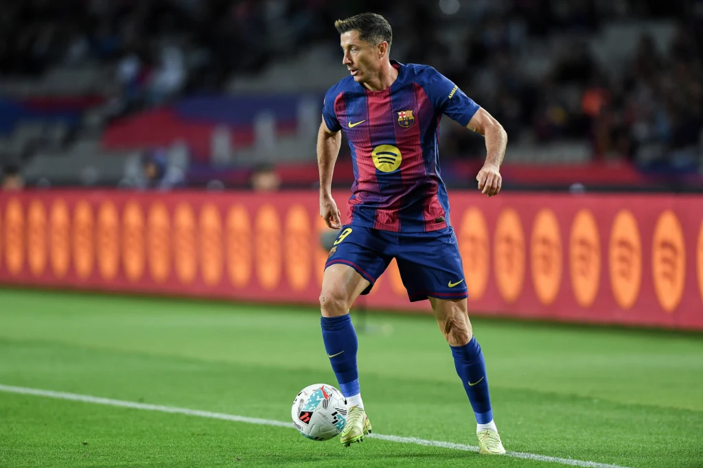 Robert Lewandowski Piłkarz w stroju FC Barcelony kontroluje piłkę na murawie boiska podczas meczu, widoczny stadion i rozmyte sylwetki kibiców w tle.