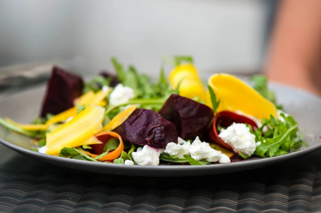 Kolorowa sałatka składająca się z rukoli, kawałków żółtej i czerwonej buraka, cienkich plastrów marchewki oraz grudek białego sera na ciemnoszarym talerzu.