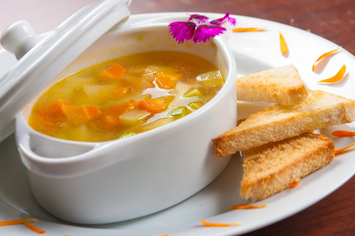 Oto jak poprawić kondycję paznokci dzięki prostym kuchennym trikom Biała miska z zupą warzywną w towarzystwie chrupiących grzanek, na talerzu ozdobionym fioletowym kwiatem i płatkami.