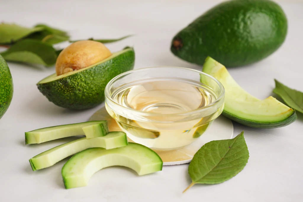 Połówki i plasterki awokado ułożone obok szklanej miseczki z olejem na białym tle, w otoczeniu zielonych liści i całych owoców awokado.