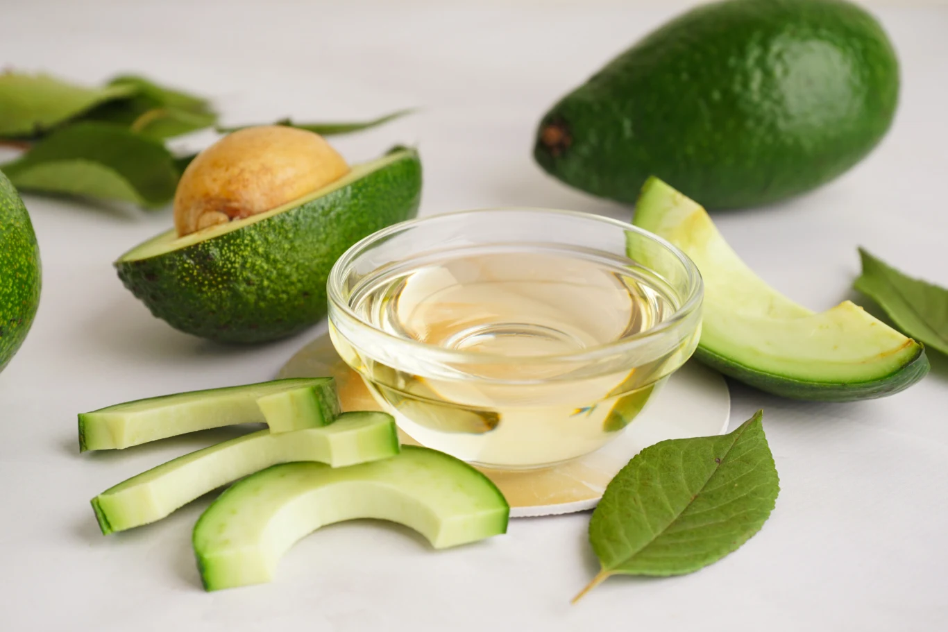 Połówki i plasterki awokado ułożone obok szklanej miseczki z olejem na białym tle, w otoczeniu zielonych liści i całych owoców awokado.