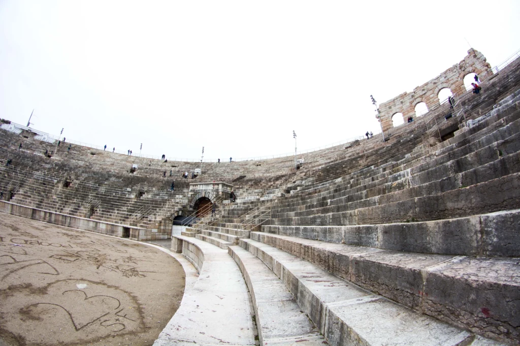 Wnętrze Amfiteatru w Weronie - areny ceremonii zamknięcia zimowych igrzysk olimpijskich 2026 Kamienne trybuny starożytnego amfiteatru z widocznymi ławkami i fragmentem areny z wyrysowanymi sercami na piasku, z częściowo zachowanymi arkadami oraz niewielką liczbą odwiedzających.