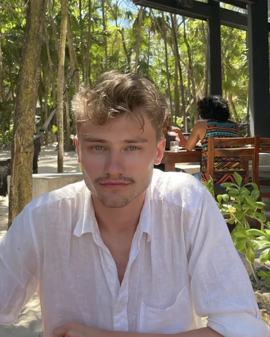 Tak wygląda obecnie 26-letni Roch - syn Kayah i Rinke Młody mężczyzna o jasnej cerze i krótkich, falowanych włosach siedzi przy stole na zewnątrz w otoczeniu tropikalnych roślin. W tle widoczna kobieta w kolorowej sukience, również siedząca przy stole. Otoczenie sugeruje relaksującą atmosferę restauracji ...