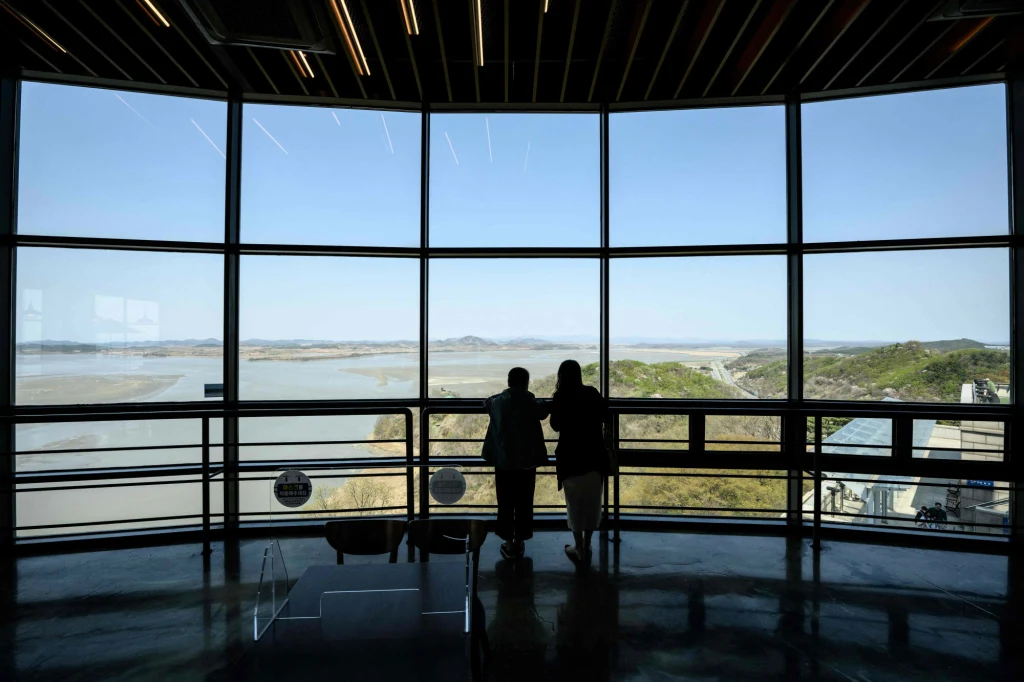 Mimo że w DMZ oficjalnie nie powinny stacjonować wojska, koreańska strefa zdemilitaryzowana to jedna z najlepiej strzeżonych granic świata Dwie osoby stoją przy dużych oknach panoramicznych, spoglądając na rozległy krajobraz rzeki oraz zielonych wzgórz rozciągających się aż po horyzont, całość oświetlona naturalnym światłem.