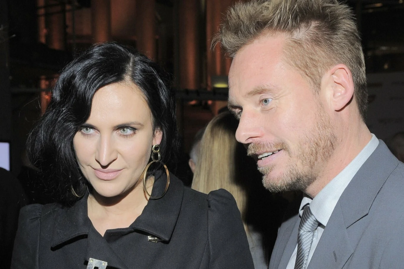 Kobieta o ciemnych włosach w dużych okrągłych kolczykach oraz mężczyzna w garniturze i jasnej koszuli, stoją obok siebie w eleganckim otoczeniu, w tle rozmyte sylwetki innych osób.