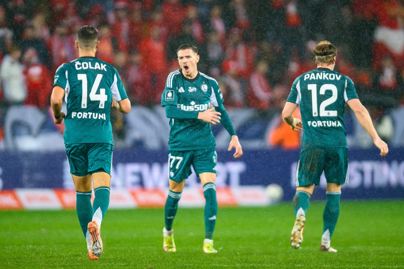 Antonio Colak, Ermal Krasniqi i Radovan Pankow w barwach Legii Warszawa Trzech piłkarzy w zielonych strojach drużyny Legia Warszawa na boisku podczas meczu, jeden zawodnik wyraża silne emocje, pozostali są zwróceni tyłem, w tle trybuny z kibicami.