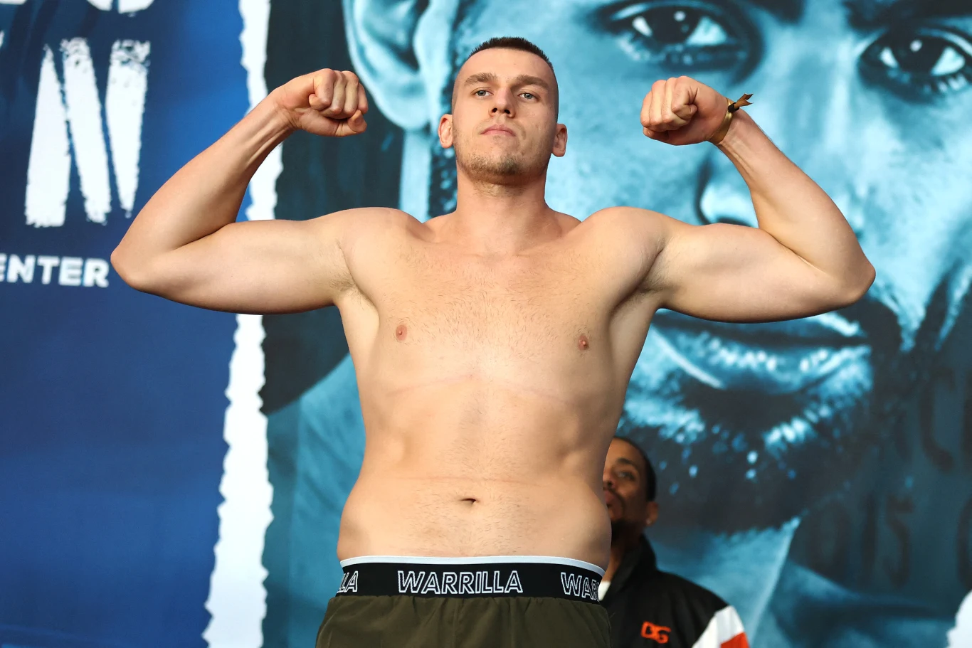 Mężczyzna z odsłoniętym torsem pozuje z napiętymi ramionami w geście pokazującym siłę, stoi na tle graficznego plakatu z twarzą innej osoby.