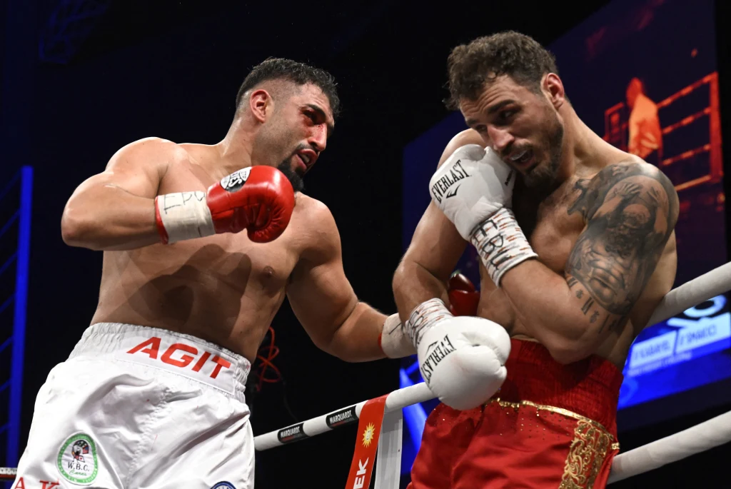 Agit Kabayel (z lewej) Dwóch bokserów podczas walki na ringu, jeden z zawodników wyprowadza silny cios w kierunku przeciwnika, który chroni się w narożniku ringu. Oświetlona arena, intensywna rywalizacja, dynamiczny moment walki sportowej.