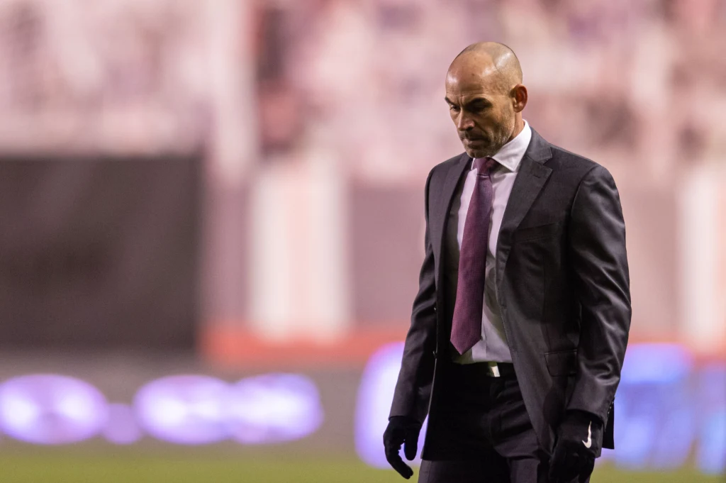 Łysy mężczyzna w garniturze z fioletowym krawatem stoi ze spuszczoną głową na tle rozmytego stadionu i widowni, sprawiając wrażenie zamyślonego lub przygnębionego.