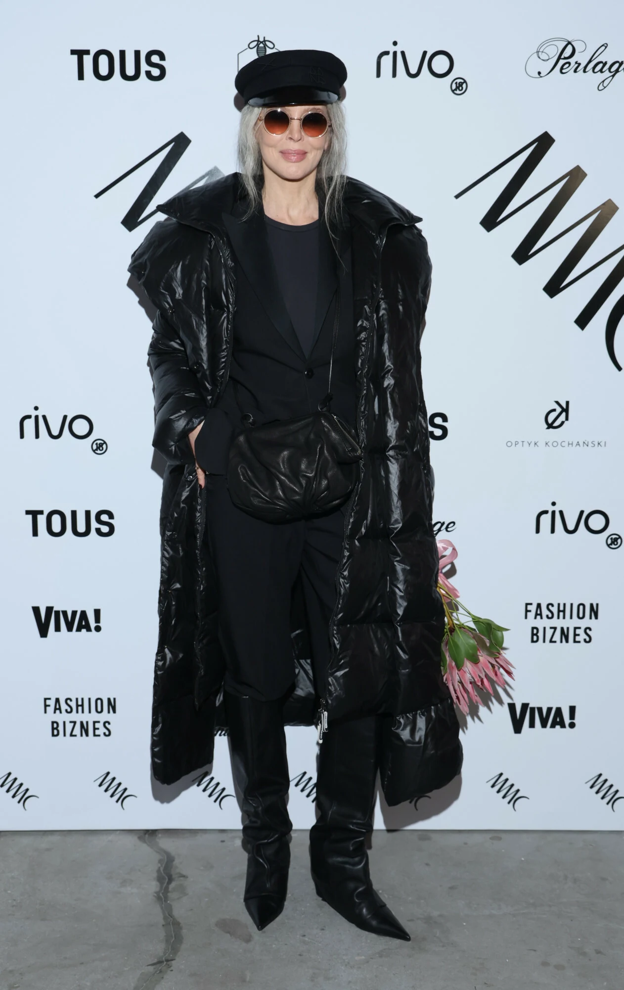 Kobieta o siwych włosach ubrana na czarno, z długim, pikowanym płaszczem, czapką z daszkiem i okrągłymi okularami przeciwsłonecznymi pozuje na tle ścianki z logo licznych marek, trzymając kwiaty.
